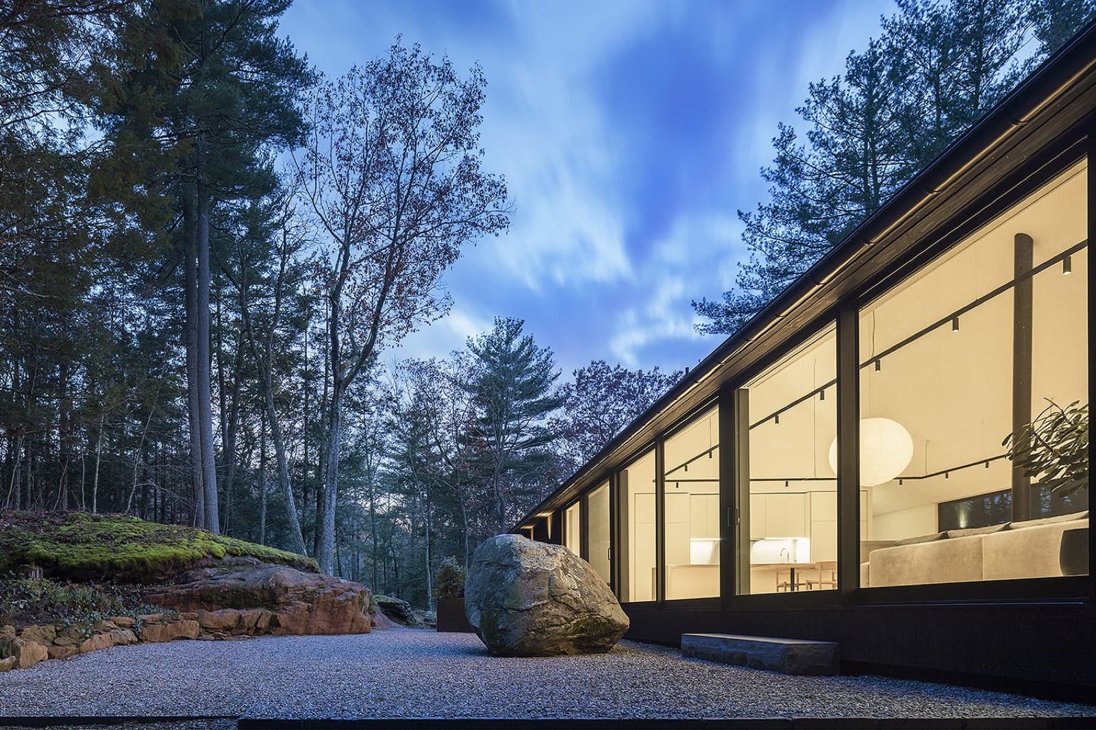 Glass-walled residence at dusk among bare trees, a single boulder in a raked gravel garden, the lit kitchen glowing within