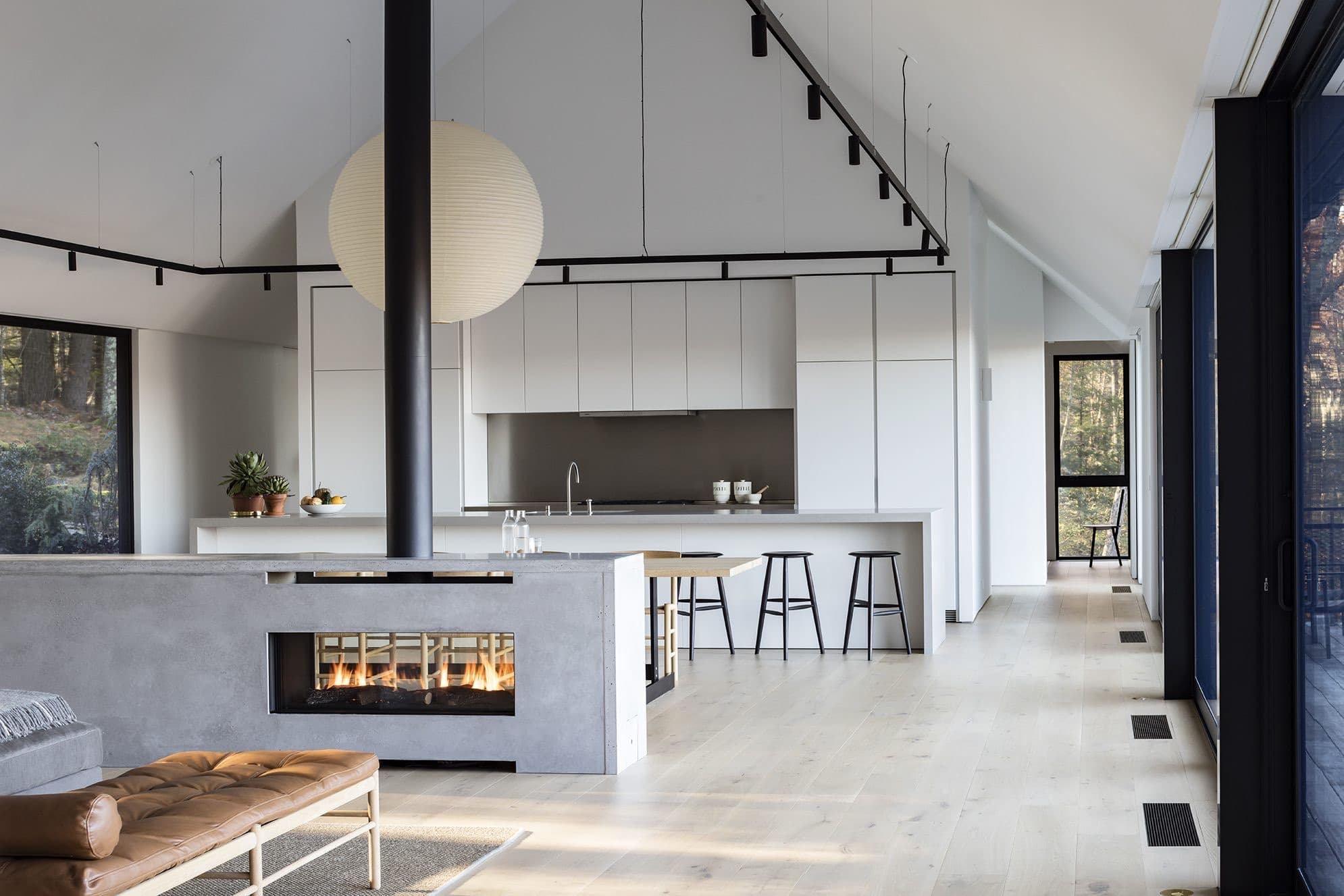 White b3 kitchen beneath a vaulted ceiling with a freestanding concrete fireplace and forest view