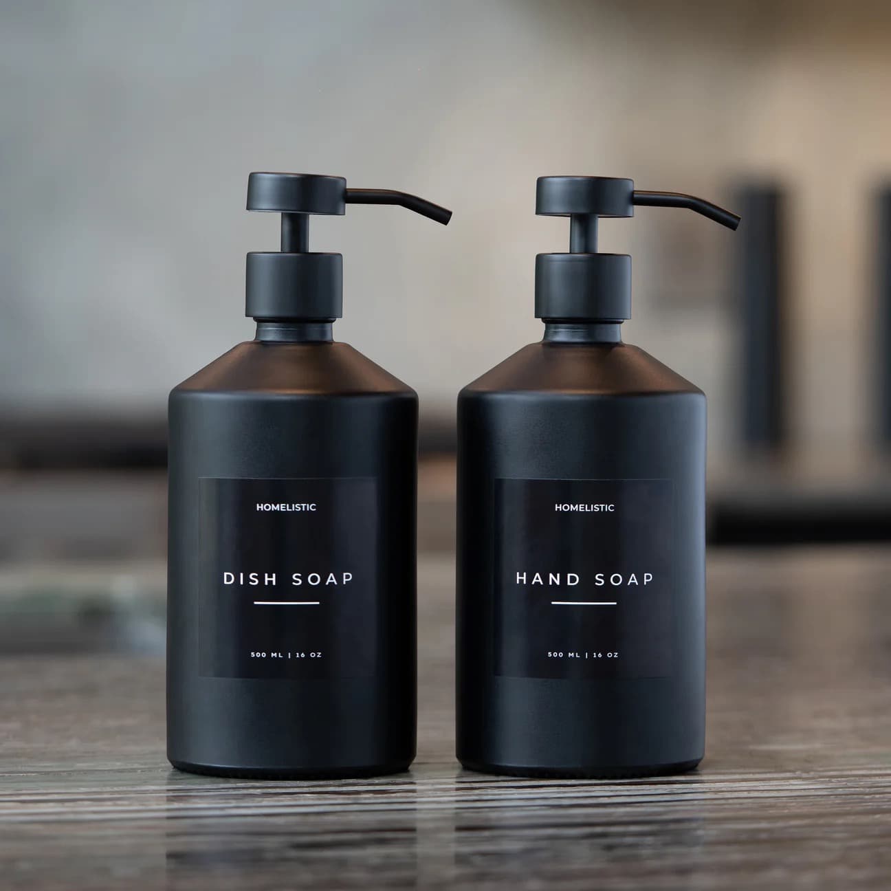 Two matte-black Homelistic dispensers in the wordmark label system — dish soap and hand soap, 500 ml / 16 oz — on a dark wooden surface