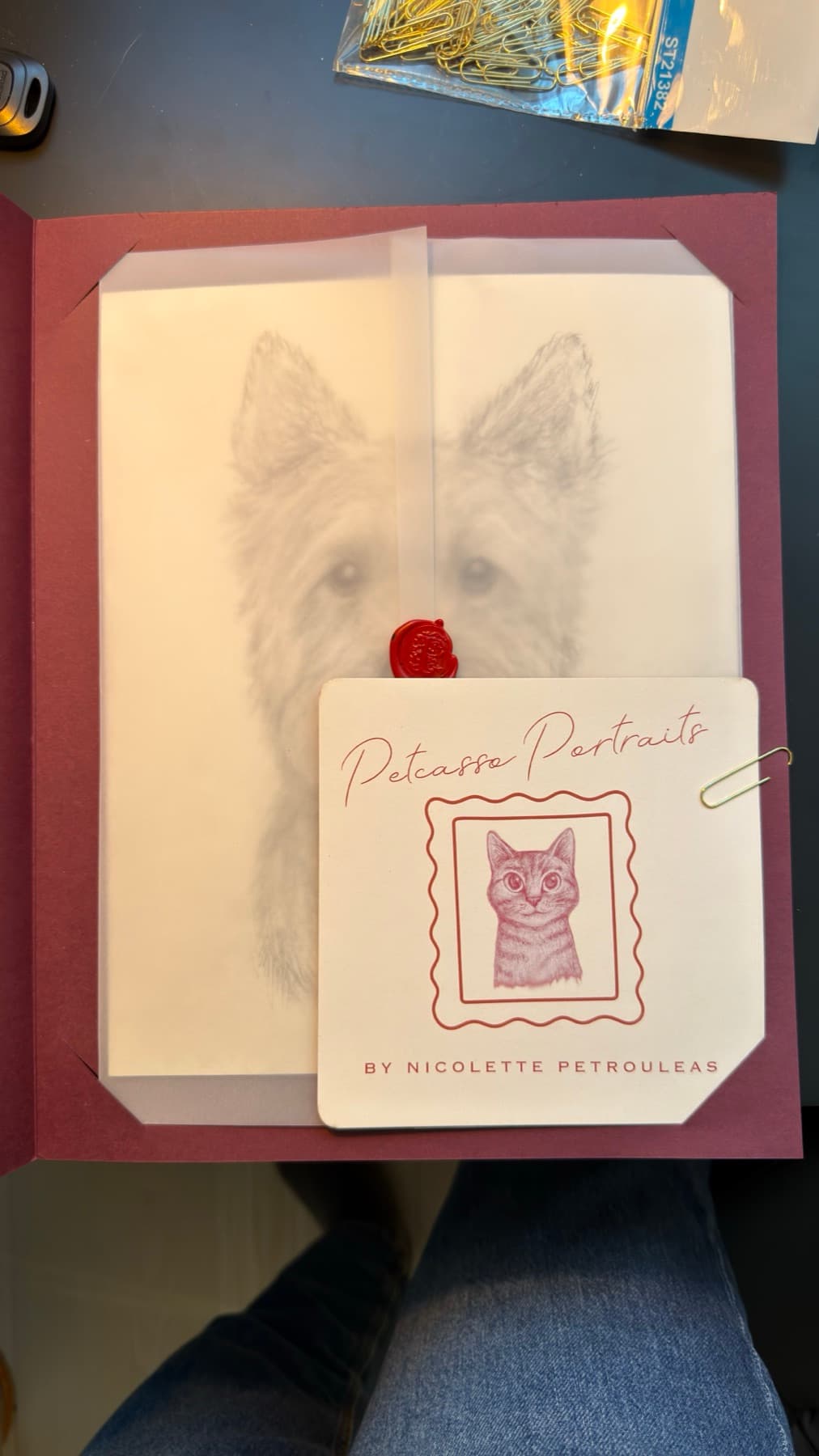An opened burgundy folio revealing a pet portrait behind vellum, sealed with red wax and a branded card