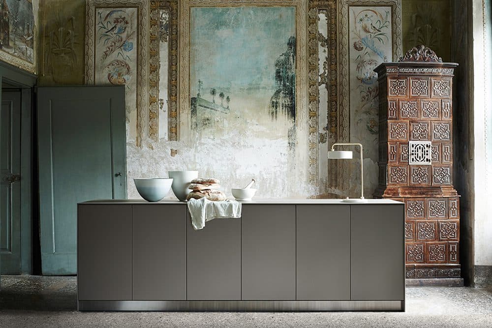 A minimalist bulthaup kitchen island set against a faded fresco wall and an antique carved stove