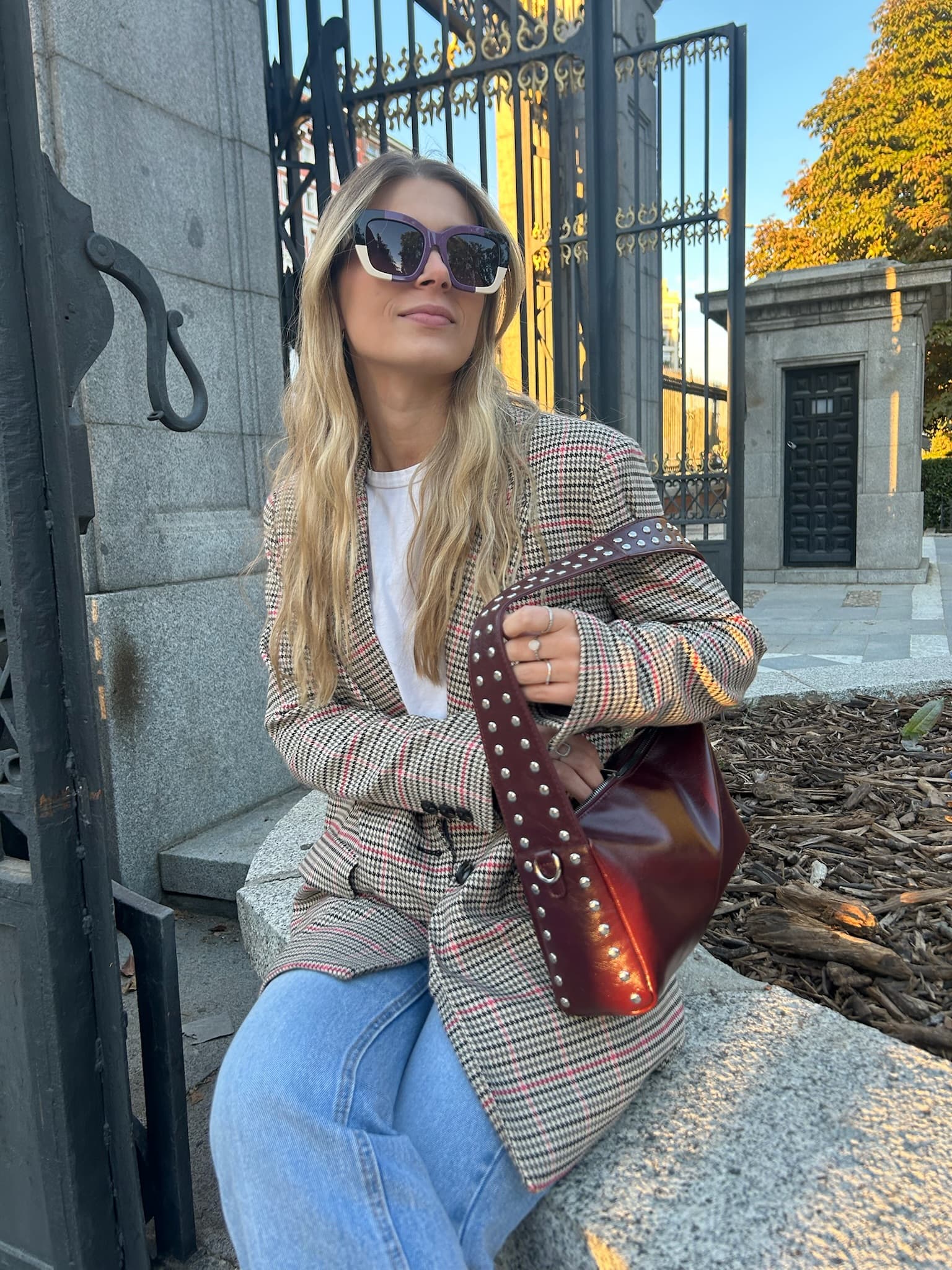 A model in a checked blazer wearing the tortoise KOHL frame, seated by an iron gate in autumn light